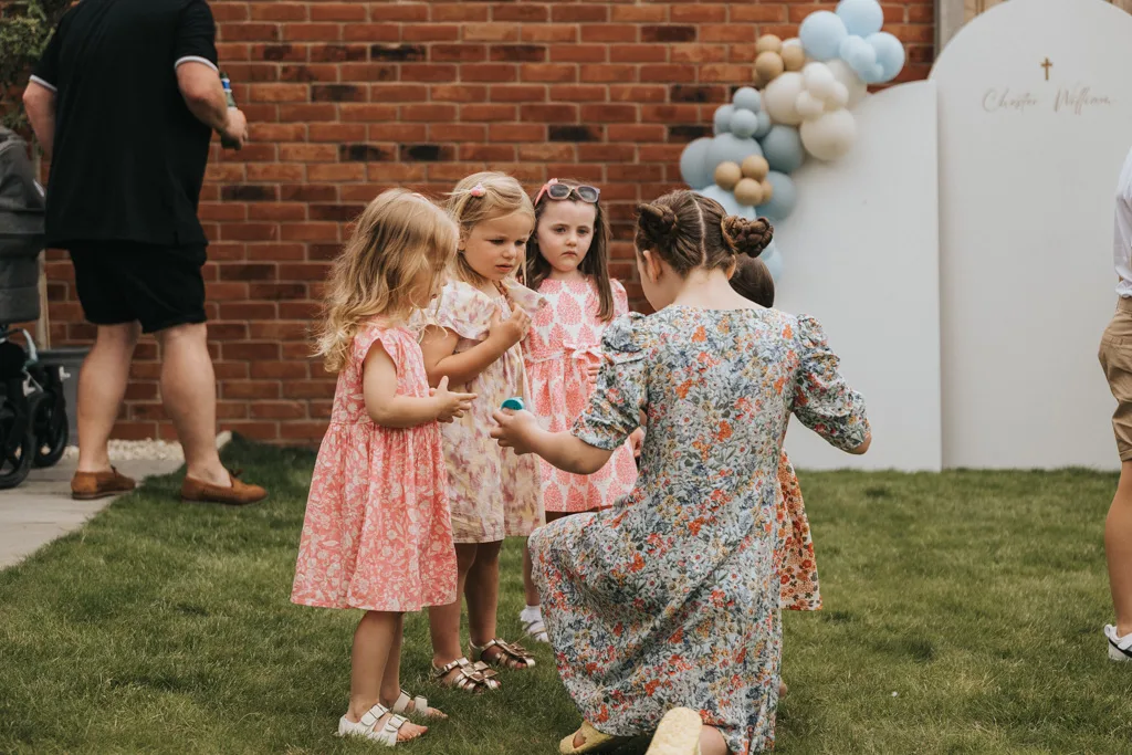 A woman kneels on the grass during a christening in Waltham, facing four young girls and handing them a small item. The girls, in floral dresses, form a semicircle. A brick wall and an arch with white, gold, and blue balloons provide the backdrop. To the left stands a man with a stroller. © Aimee Lince Photography - Wedding photographer in Lincolnshire, Yorkshire & Nottinghamshire