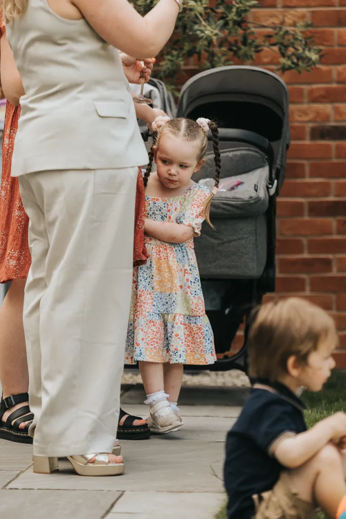 A young girl in a colorful, floral dress stands with arms crossed, looking down, perhaps pondering the day's festivities in Grimsby. She has braided hair and is next to a woman in white pants, while a stroller can be seen behind them. A toddler in a navy shirt sits on the ground, blurred in the foreground. They're outside on a paved area. © Aimee Lince Photography - Wedding photographer in Lincolnshire, Yorkshire & Nottinghamshire