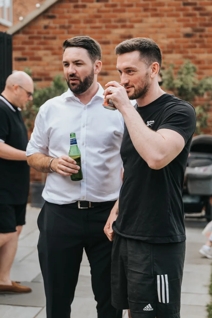 In a casual outdoor setting in Waltham, two men stand relaxed. One, wearing a white shirt and dark pants, holds a green bottle, while the other in a black t-shirt and shorts sips from a can. Behind them, another person stands near brick walls adorned with greenery. © Aimee Lince Photography - Wedding photographer in Lincolnshire, Yorkshire & Nottinghamshire