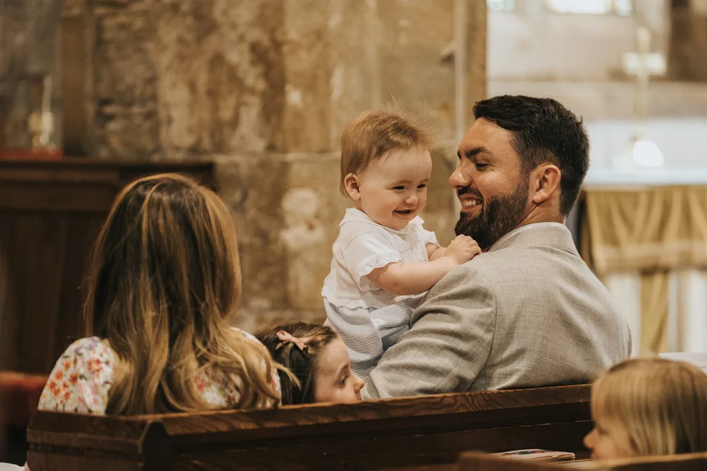 A cheerful baby in white clothes is held by a bearded man in a light gray suit, celebrating a christening inside a church. The man smiles warmly at the baby. A woman with long hair sits beside them, partially visible against the rustic stone walls and wooden bench. © Aimee Lince Photography - Wedding photographer in Lincolnshire, Yorkshire & Nottinghamshire