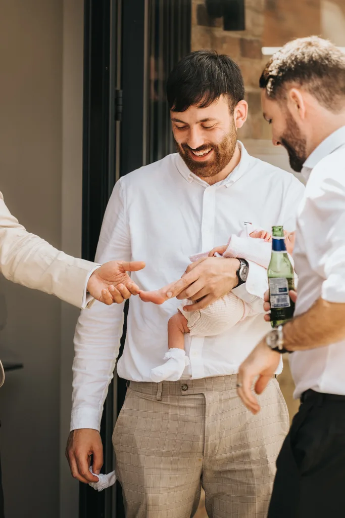 A man in a white shirt and plaid pants holds a baby with a proud smile, presumably at a christening. Two others, also in white shirts, join him—one clutching a beer bottle—as another hand reaches toward the baby. They're gathered outside near a building in Waltham. © Aimee Lince Photography - Wedding photographer in Lincolnshire, Yorkshire & Nottinghamshire
