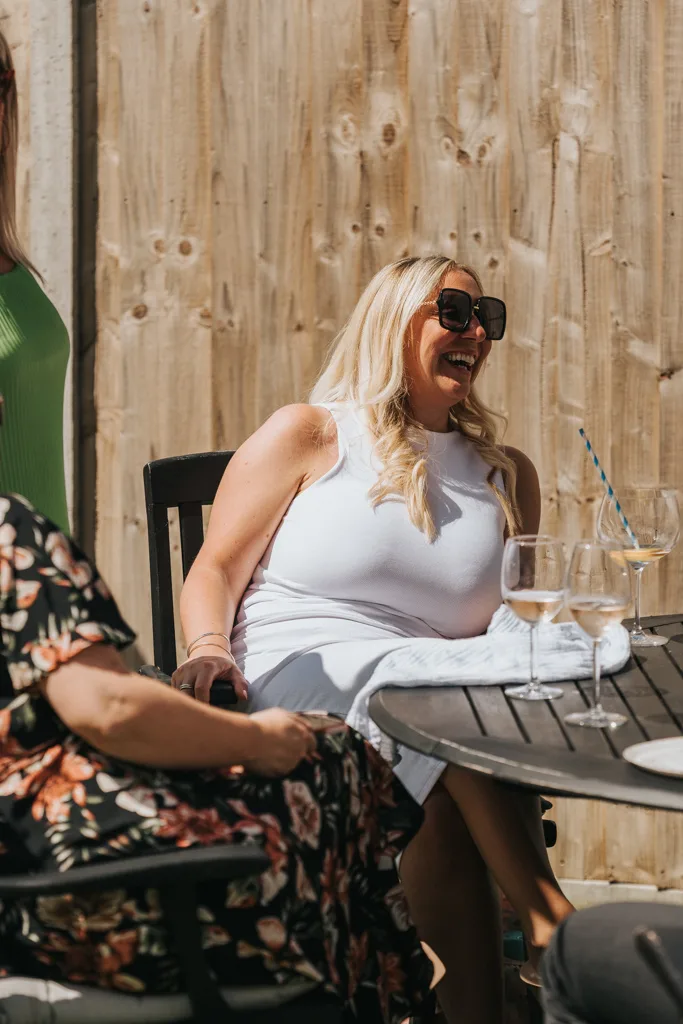 A woman with long blonde hair and large sunglasses is laughing while sitting at an outdoor table in Waltham. She wears a white sleeveless top and has a napkin on her lap. The table holds wine glasses, with a wooden fence in the background. Another person in floral attire is partially visible. © Aimee Lince Photography - Wedding photographer in Lincolnshire, Yorkshire & Nottinghamshire