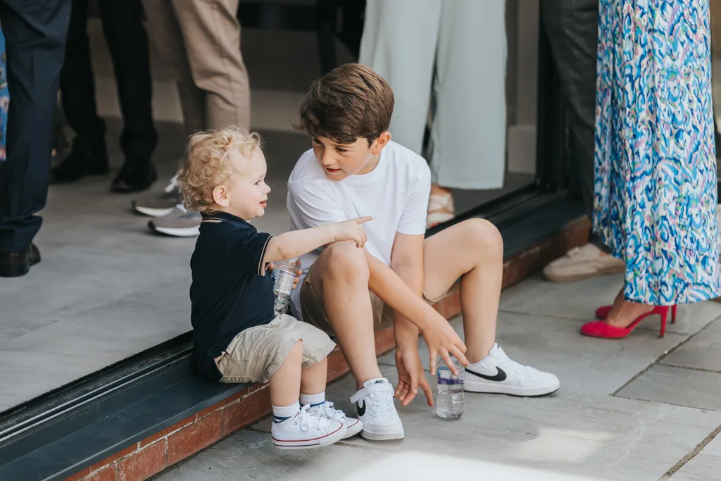 Two young boys sit on a doorstep in Waltham. The older boy, wearing a white t-shirt and shorts, holds a water bottle and looks at the younger one in a black shirt and khaki shorts. They are deep in conversation about the christening they just attended as adults stand nearby. © Aimee Lince Photography - Wedding photographer in Lincolnshire, Yorkshire & Nottinghamshire