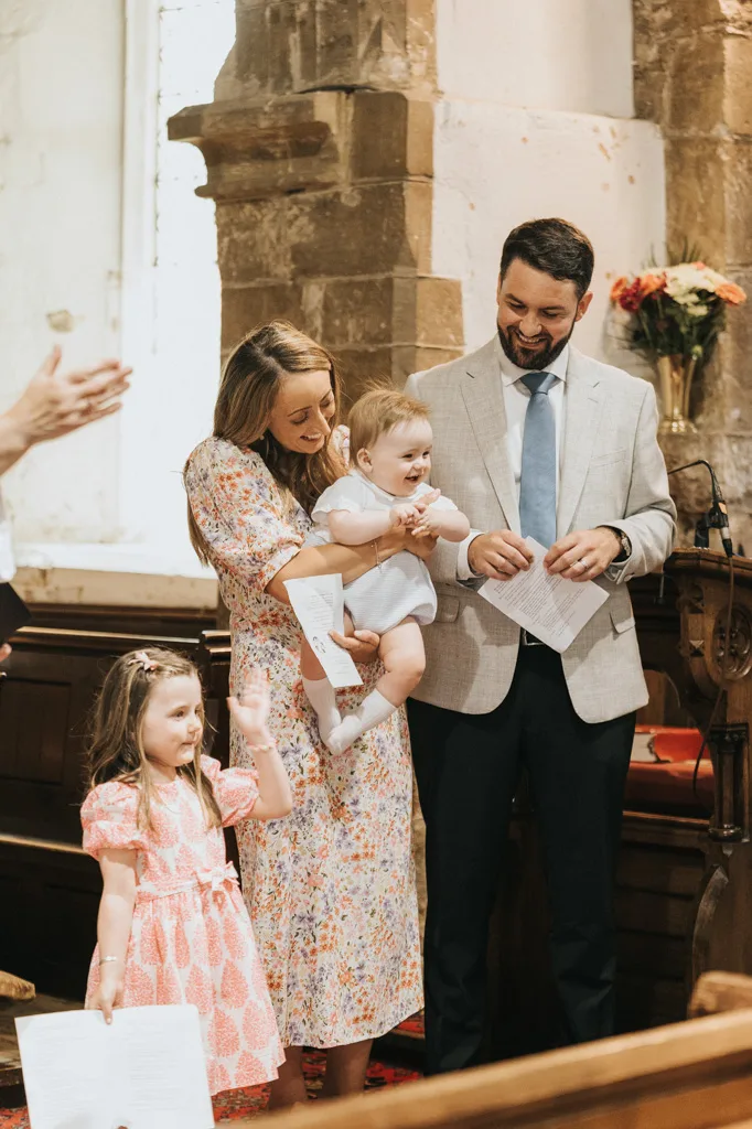 A family stands indoors, beaming. A woman in a floral dress holds a baby, freshly christened. A man in a light gray suit and blue tie stands next to her, holding the certificate. A young girl in a pink dress stands prominently. They're in a room with stone walls, perhaps in Waltham, and flowers adorn the background. © Aimee Lince Photography - Wedding photographer in Lincolnshire, Yorkshire & Nottinghamshire