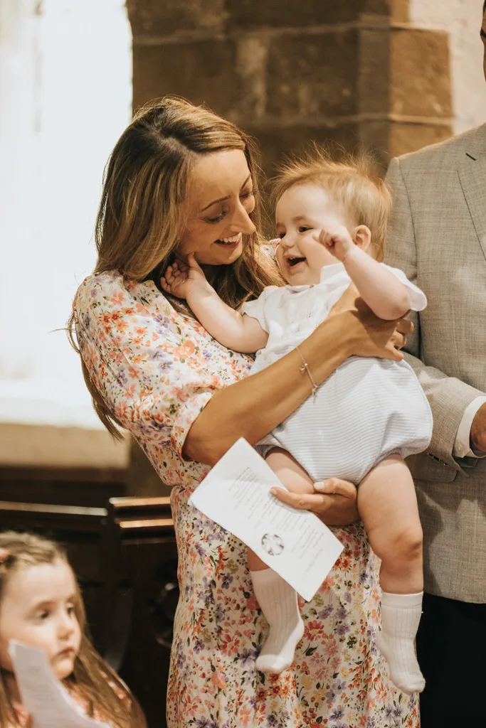 In a warmly lit church, a woman in a floral dress joyfully cradles a smiling baby dressed in white and blue, celebrating their christening. The baby's eyes shine as they gaze up at her. Nearby, another child with a serious expression peeks into the scene, adding to the Grimsby gathering's charm. © Aimee Lince Photography - Wedding photographer in Lincolnshire, Yorkshire & Nottinghamshire