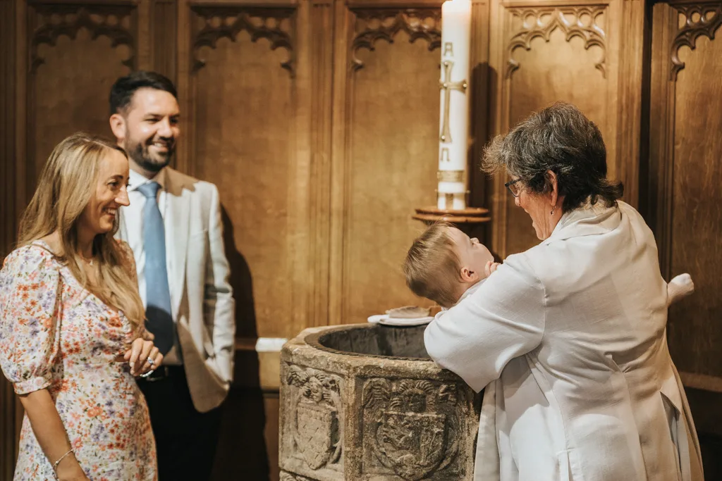 A baby is being christened by a woman in a white robe over an ornate stone font. A tall candle glows warmly in the background. A smiling woman in a floral dress and a man in a suit look on, adding joy to the ceremony. The wooden paneling enhances the inviting ambiance of this Waltham church scene. © Aimee Lince Photography - Wedding photographer in Lincolnshire, Yorkshire & Nottinghamshire