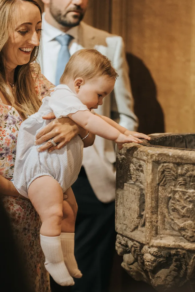 A smiling woman in a floral dress holds a baby dressed in a white shirt and blue romper, reaching towards an ornate stone baptismal font during the christening. A man in a light suit and blue tie stands in the background, capturing the sacred moment inside the Grimsby church. © Aimee Lince Photography - Wedding photographer in Lincolnshire, Yorkshire & Nottinghamshire