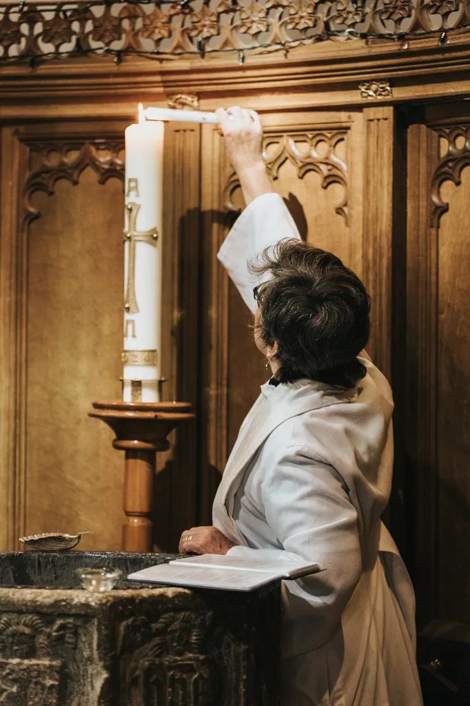 A person in a white robe lights a tall candle in a carved wooden church setting in Waltham. The candle, marked with an Alpha and Omega symbol, stands on a pedestal beside a decorative stone font used for christenings. The warm glow illuminates the intricate carvings and wooden paneling behind. © Aimee Lince Photography - Wedding photographer in Lincolnshire, Yorkshire & Nottinghamshire