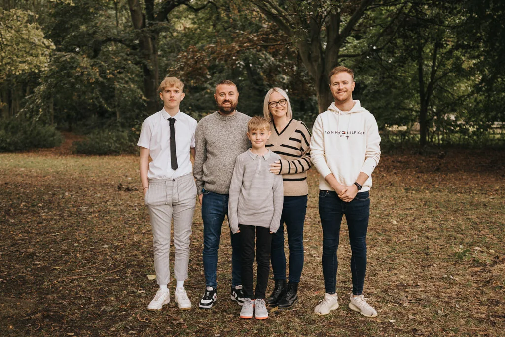 Amid the leafy backdrop of Grimsby, a family of five stands together for a casual photoshoot. Arranged in a row, the parents are centered with a child in front and two young men flanking them, all sharing soft smiles among the towering trees. © Aimee Lince Photography - Wedding photographer in Lincolnshire, Yorkshire & Nottinghamshire