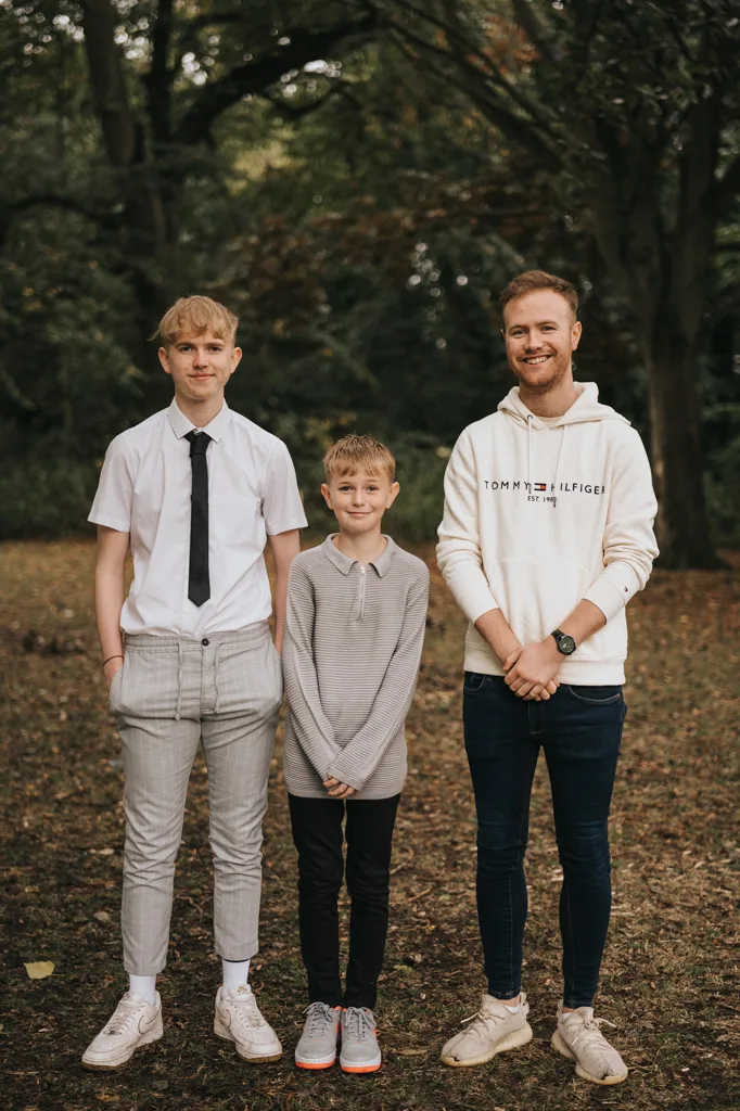 In a serene wooded area near Grimsby, three people gather for a cheerful photoshoot. The one on the left dons a white shirt and tie with checked pants, the middle sports a light gray sweater, and the right is in a white hoodie with jeans. Surrounded by trees and fallen leaves, they all smile brightly. © Aimee Lince Photography - Wedding photographer in Lincolnshire, Yorkshire & Nottinghamshire