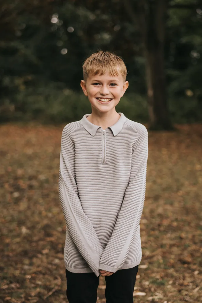 A young boy stands smiling outdoors in a park, captured by a talented photographer. He has light brown hair and wears a light gray, long-sleeved sweater with a collar and buttons. The blurred background reveals trees in Grimsby and dry leaves, creating a relaxed and cheerful family atmosphere. © Aimee Lince Photography - Wedding photographer in Lincolnshire, Yorkshire & Nottinghamshire