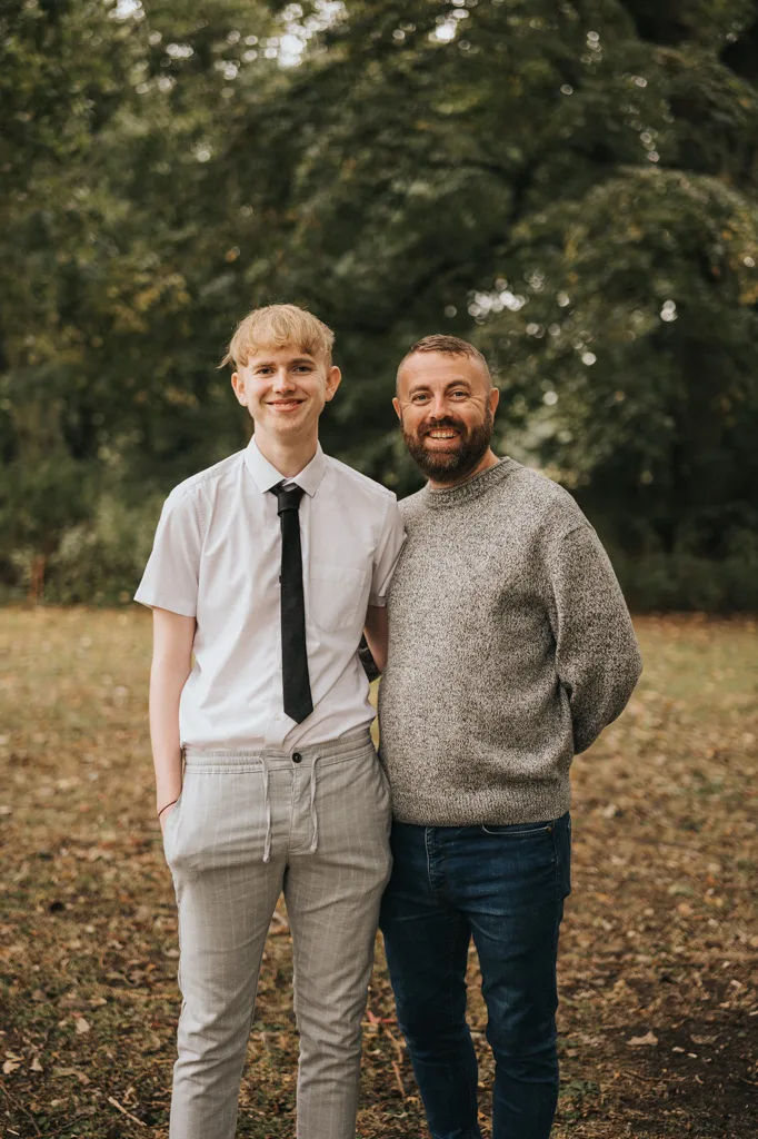 During a photoshoot in Grimsby's picturesque outdoors, a young man in a white shirt and tie stands beside an older man in a gray sweater and jeans. Both are smiling warmly at the camera, framed by lush green trees. The young man's hands rest in his pockets while the older man has an arm around him. © Aimee Lince Photography - Wedding photographer in Lincolnshire, Yorkshire & Nottinghamshire