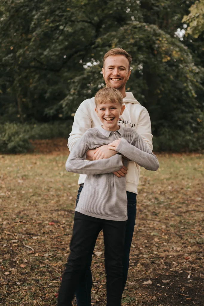 A smiling man in a white hoodie hugs a laughing boy in a gray sweater from behind during their family photoshoot. They stand outdoors on a grassy area with lush, green trees as their backdrop. The ground is scattered with fallen leaves, and both appear joyful and relaxed. © Aimee Lince Photography - Wedding photographer in Lincolnshire, Yorkshire & Nottinghamshire