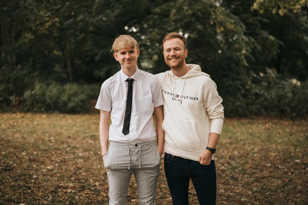 Two young men stand outdoors on a grassy, leaf-strewn area during a photoshoot in Grimsby. The first wears a white shirt, black tie, and gray pants. The second sports a light-colored hoodie with "Tommy Hilfiger" on it and dark pants. Both are smiling, with trees in the background. © Aimee Lince Photography - Wedding photographer in Lincolnshire, Yorkshire & Nottinghamshire