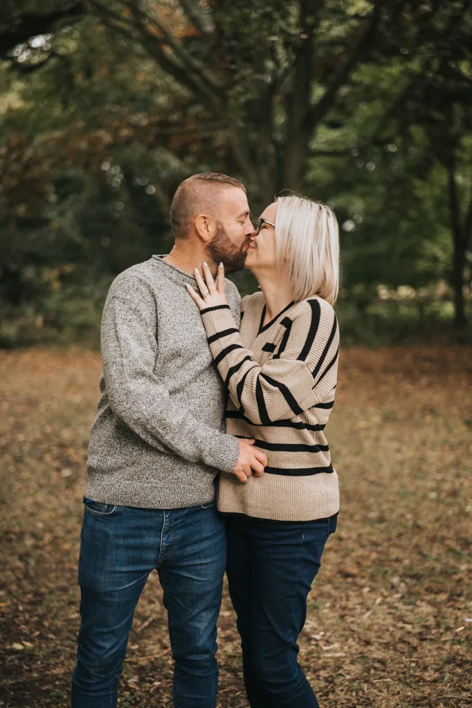A couple shares a kiss in a park, captured perfectly by a photographer. The man wears a gray sweater and jeans, while the woman dons a striped sweater and jeans. Surrounded by trees and fallen leaves, this romantic autumn scene could be part of any cherished family photoshoot. © Aimee Lince Photography - Wedding photographer in Lincolnshire, Yorkshire & Nottinghamshire