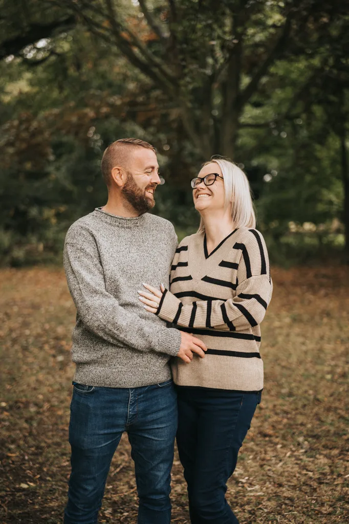 A smiling couple stands in a wooded area with autumn leaves underfoot, captured perfectly by the photographer. The man, in a gray sweater and jeans, has his arm around the woman. She wears glasses, a beige sweater with black stripes, and jeans. They look at each other affectionately amidst green trees—a timeless photoshoot portrait. © Aimee Lince Photography - Wedding photographer in Lincolnshire, Yorkshire & Nottinghamshire