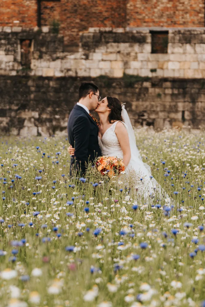 A bride in a white gown and veil kisses a groom in a dark suit amid wildflowers, including cornflowers and daisies. The groom holds a bouquet of orange flowers. A rustic stone wall adds historical charm to the romantic scene, perfect for any wedding portfolio showcasing timeless love. © Aimee Lince Photography - Wedding photographer in Lincolnshire, Yorkshire & Nottinghamshire