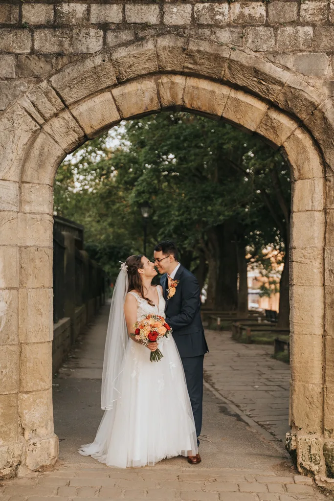 A bride in a white gown and veil holding a bouquet stands under a stone archway with her groom in a dark suit. Smiling and close, they’re framed by trees and a cobblestone path. Capturing this moment adds timeless beauty to any wedding portfolio. The groom wears glasses and a boutonniere. © Aimee Lince Photography - Wedding photographer in Lincolnshire, Yorkshire & Nottinghamshire