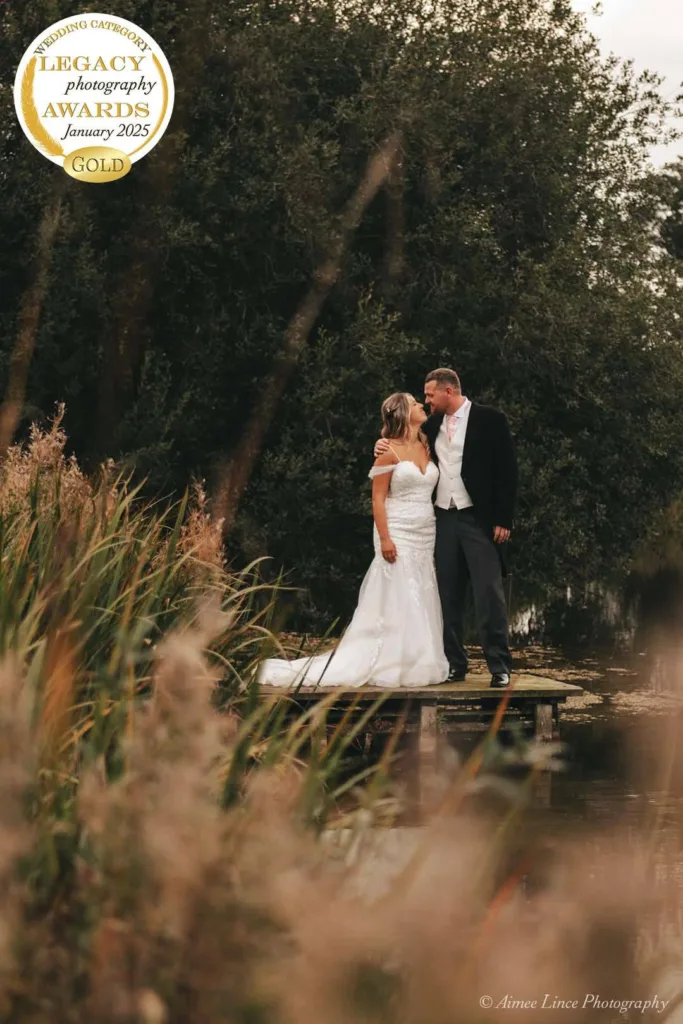 A bride in a white gown and a groom in a tuxedo share a kiss on a small wooden dock surrounded by greenery. Tall grasses are in the foreground. A gold medal in the top left corner reads “Legacy Photography Awards, January 2025, Winner.” Photo by Aimée Lince Photography. © Aimee Lince Photography - Wedding photographer in Lincolnshire, Yorkshire & Nottinghamshire