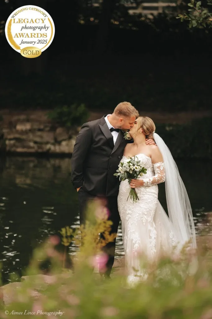 A bride and groom kiss by a tranquil river, captured in exquisite wedding photography. She wears a long, white lace gown with a veil, holding a bouquet, while the groom stands beside her in a black tuxedo. A "Legacy Photography Awards Gold" medal for January 2025 graces the corner. © Aimee Lince Photography - Wedding photographer in Lincolnshire, Yorkshire & Nottinghamshire