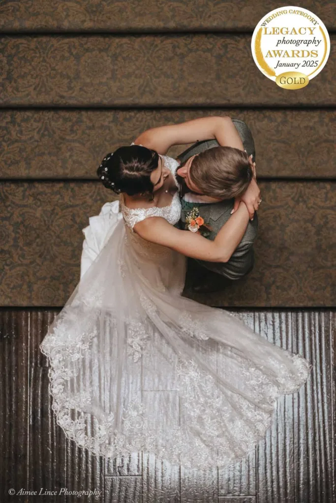 Bride and groom dance on a dark wooden floor, embracing each other. The bride's lace gown fans out gracefully. A gold medal for "Legacy Photography Awards January 2025" shines in the top right corner. Photo credits to Aimee Lince Photography, renowned in wedding photography, are at the bottom. © Aimee Lince Photography - Wedding photographer in Lincolnshire, Yorkshire & Nottinghamshire