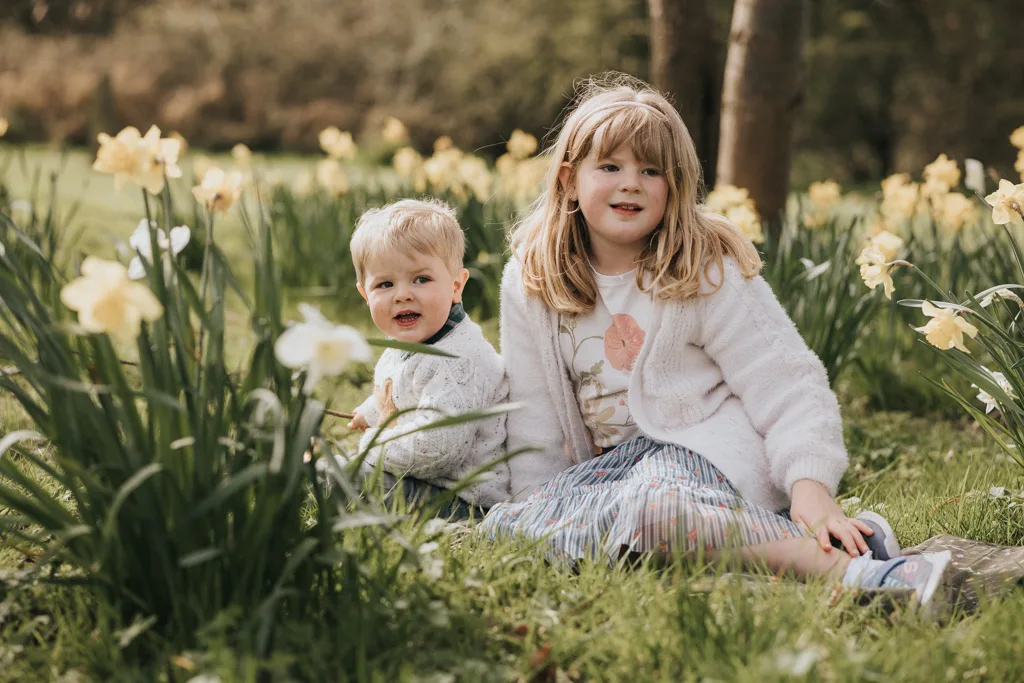 A young girl and toddler sit on the grass among blooming daffodils, celebrating the joy of Easter. The girl wears a white cardigan and colorful skirt, while the toddler sports a light sweater. Both have light hair and smiles, framed by a tree trunk and soft-focus greenery in the background. © Aimee Lince Photography - Wedding photographer in Lincolnshire, Yorkshire & Nottinghamshire