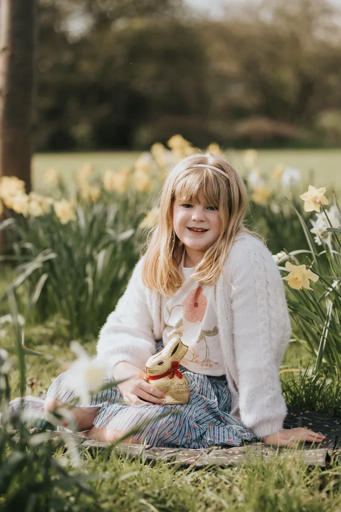 A young girl with blonde hair sits on a blanket in a garden surrounded by daffodils. Celebrating Easter, she wears a white cardigan and a floral dress, smiling at the camera while holding a chocolate bunny wrapped in gold foil. It's a sunny day with greenery in the background. © Aimee Lince Photography - Wedding photographer in Lincolnshire, Yorkshire & Nottinghamshire