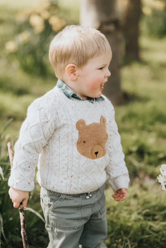 A young child with blonde hair stands outdoors on grassy terrain, celebrating the Easter season. Wearing a white sweater with a bear design and green pants, the child holds a stick in one hand. Yellow flowers and trees are visible in the blurred background, suggesting a spring or summer setting. © Aimee Lince Photography - Wedding photographer in Lincolnshire, Yorkshire & Nottinghamshire