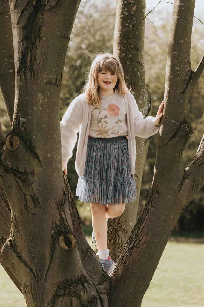 A young girl, dressed for Easter in a white cardigan and floral shirt, is climbing a tree. She sports a blue pleated skirt and sneakers, her hair down as she smiles. The tree's thick branches and textured bark add charm to the grassy backdrop with its blurred foliage. © Aimee Lince Photography - Wedding photographer in Lincolnshire, Yorkshire & Nottinghamshire