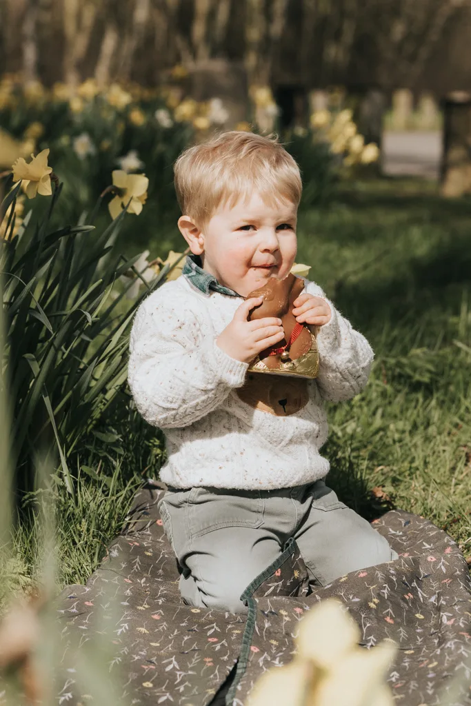 A toddler with blond hair sits on a patterned blanket in a grassy area, surrounded by blooming yellow flowers. Clad in a white sweater and gray pants, they happily bite into an Easter chocolate bunny, with some chocolate smeared on their face. The sunlit scene is pure joy. © Aimee Lince Photography - Wedding photographer in Lincolnshire, Yorkshire & Nottinghamshire