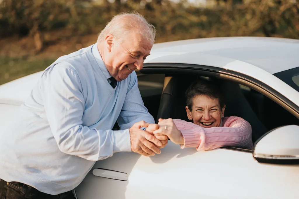 Elderly man smiling, holding hands with an elderly woman in a white car, their joyful engagement evident. She's in a pink sweater, smiles warmly through the open window. The blurred background hints at the charm of Cleethorpes' outdoor setting. © Aimee Lince Photography - Wedding photographer in Lincolnshire, Yorkshire & Nottinghamshire