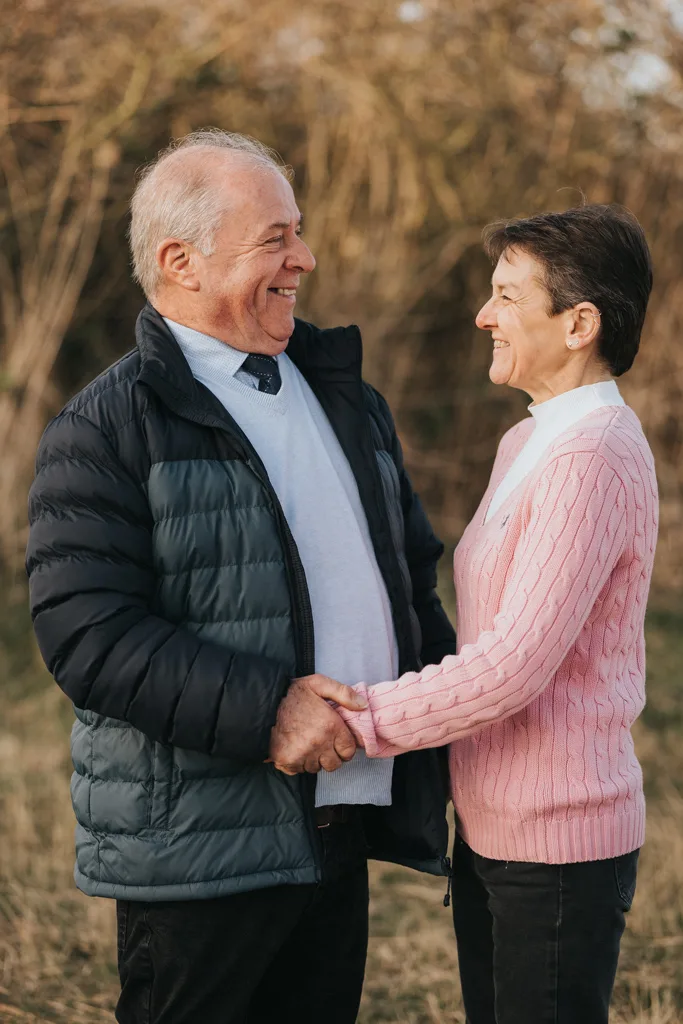 An elderly man and woman stand outside in Cleethorpes, smiling at each other and holding hands. The man wears a black puffer jacket over a white shirt, while the woman sports a pink cable-knit sweater. Their engagement radiates joy against the backdrop of tall grass and soft sunlight. © Aimee Lince Photography - Wedding photographer in Lincolnshire, Yorkshire & Nottinghamshire