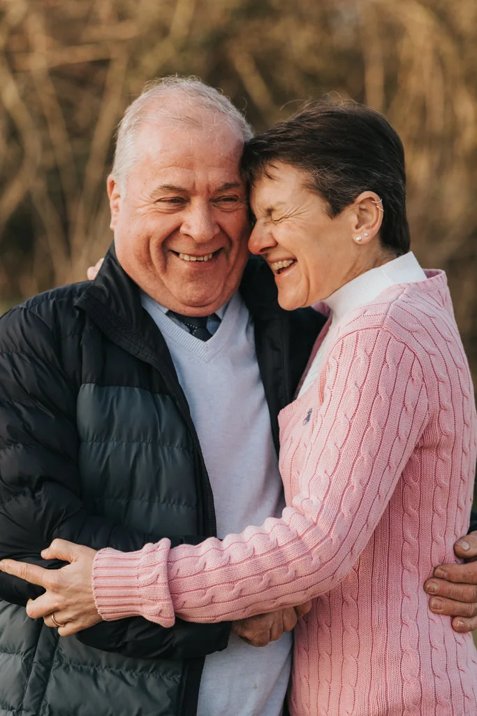 An elderly couple stands outdoors in Cleethorpes, warmly embracing. The man, wearing a black jacket and light sweater, is smiling. The woman, in a pink cable-knit sweater, laughs with her eyes closed, leaning into him. The background is softly blurred, suggesting a natural setting. © Aimee Lince Photography - Wedding photographer in Lincolnshire, Yorkshire & Nottinghamshire