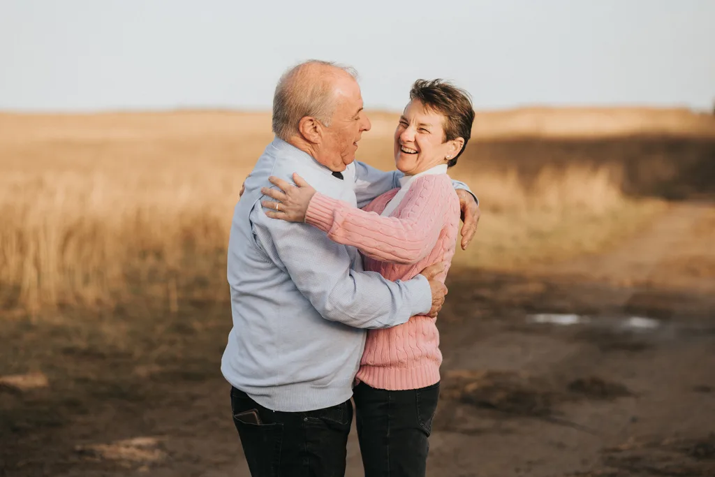 An elderly couple, recently celebrating their engagement, embrace and smile at each other in an open field. The man dons a light blue sweater while the woman wears pink. Golden grasses sway under a clear Cleethorpes sky, with soil paths weaving warmth and affection beneath them. © Aimee Lince Photography - Wedding photographer in Lincolnshire, Yorkshire & Nottinghamshire
