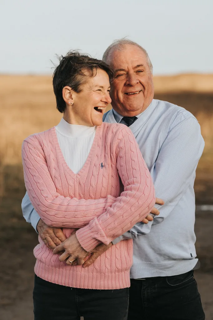 An older couple stands outdoors on a sunny day, radiating joy reminiscent of an engagement photo. The woman, in a pink sweater over a white top, laughs while looking to the side. The man, in a light blue sweater, smiles and embraces her against the blurred landscape near Cleethorpes' brown grass. © Aimee Lince Photography - Wedding photographer in Lincolnshire, Yorkshire & Nottinghamshire