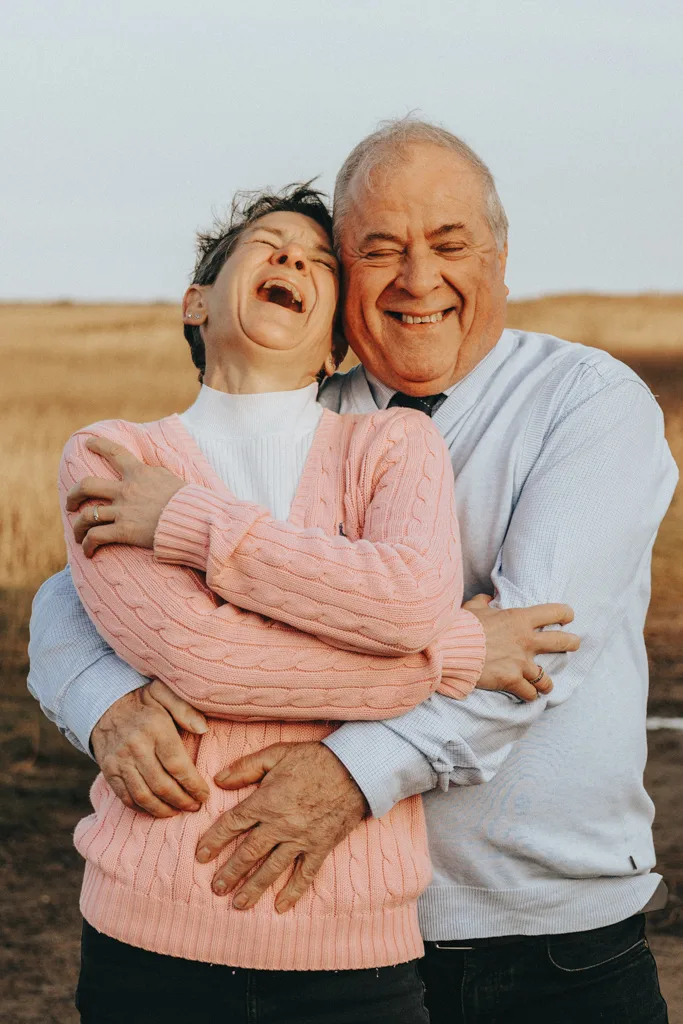 An older couple embracing and laughing in a field near Cleethorpes. The woman, in a pink sweater and white turtleneck, leans back joyfully into the man in a light blue shirt. They stand closely, his arms wrapping around her, against a backdrop of golden grass under a pale sky. © Aimee Lince Photography - Wedding photographer in Lincolnshire, Yorkshire & Nottinghamshire