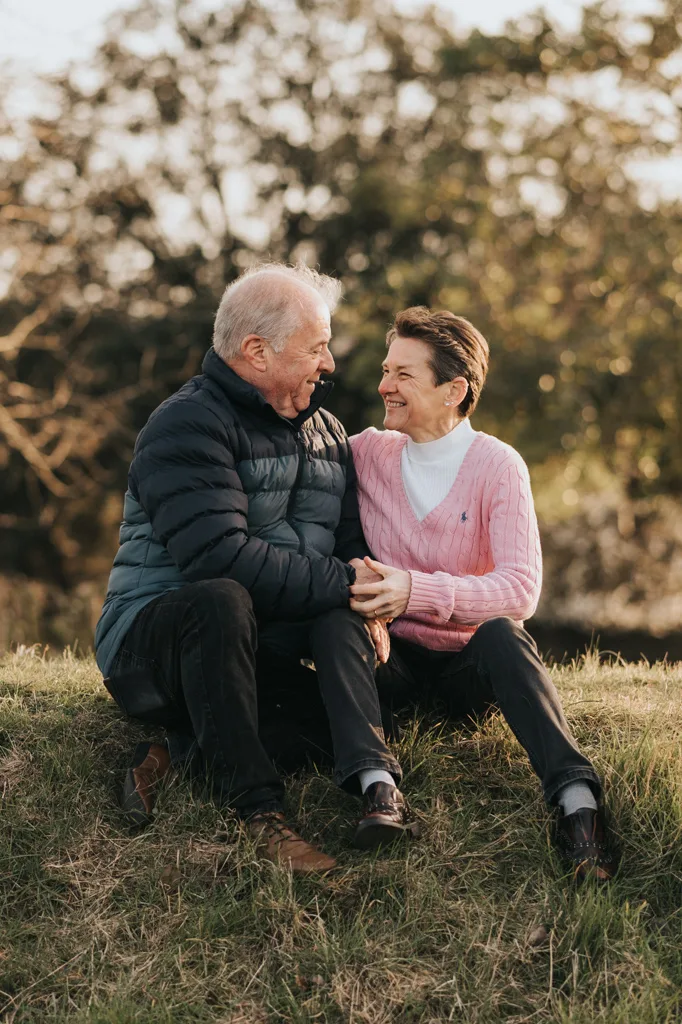 An older couple sits on the grass in Cleethorpes, smiling at each other. The man wears a dark puffer jacket and pants, while the woman dons a pink sweater over a white shirt and dark pants. Holding hands, they bask in soft sunlight with blurred trees behind them, celebrating their engagement. © Aimee Lince Photography - Wedding photographer in Lincolnshire, Yorkshire & Nottinghamshire