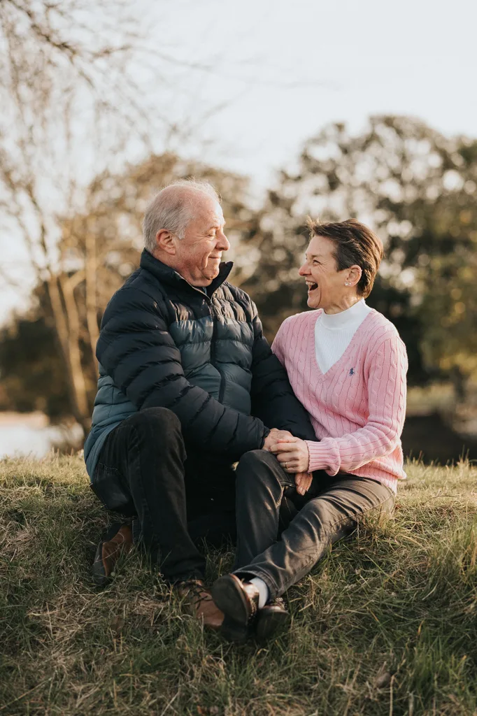 An elderly man in a puffy jacket and an elderly woman in a pink sweater sit on the grass, holding hands and laughing. Their joy is evident against the serene setting of Cleethorpes, with trees and a calm lake behind them, suggesting an engagement amidst cool, peaceful weather. © Aimee Lince Photography - Wedding photographer in Lincolnshire, Yorkshire & Nottinghamshire