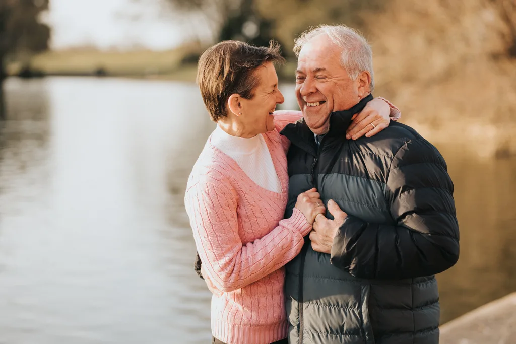 An elderly couple stands by a serene lakeside in Cleethorpes, smiling warmly at each other. The woman, wearing a pink sweater, clasps the man's jacket, while he, in a puffy black jacket, holds her arm gently. The background features a blurred view of trees and water under a clear sky. © Aimee Lince Photography - Wedding photographer in Lincolnshire, Yorkshire & Nottinghamshire