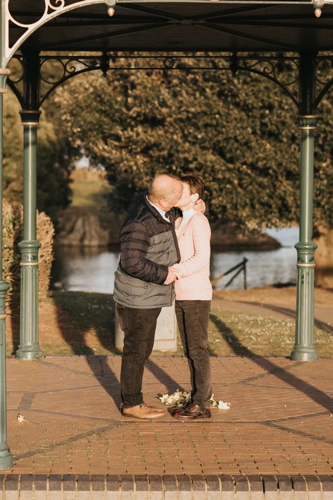 A couple shares an engagement kiss under a gazebo with ornate metalwork, enveloped by lush greenery. The man in a blue and black jacket, the woman in pink, stand on paved ground. In the backdrop of Cleethorpes, a river and trees add to the romantic outdoor setting. © Aimee Lince Photography - Wedding photographer in Lincolnshire, Yorkshire & Nottinghamshire