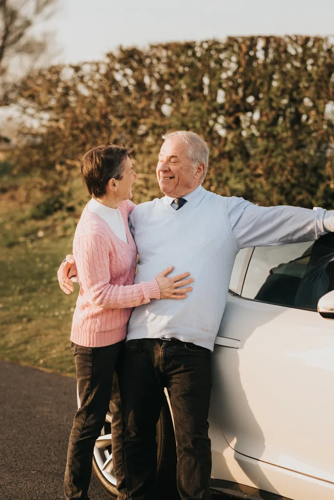 An older couple stands beside a silver car on a sunny day in Cleethorpes. Both are smiling and embracing; the woman wears a pink sweater and the man a light blue sweater. Green bushes and grass are visible in the background. © Aimee Lince Photography - Wedding photographer in Lincolnshire, Yorkshire & Nottinghamshire