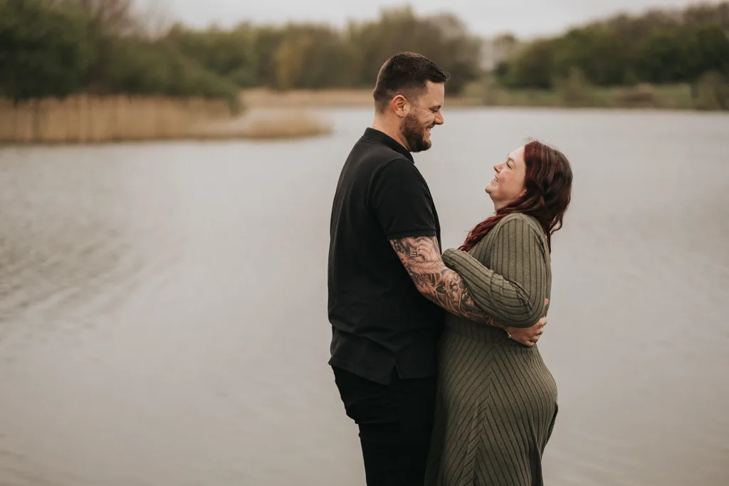 Emily & Billy Engagement Couples Phtooshoot in Cleethorpes Country Park 11 of 45 © Aimee Lince Photography - 23.04.2025 A tattooed man embraces his partner in a green dress by the water, both smiling tenderly at each other. This candid engagement photography captures couples in a natural setting, with trees and reeds softly blurred in the background. © Aimee Lince Photography - Wedding photographer in Lincolnshire, Yorkshire & Nottinghamshire