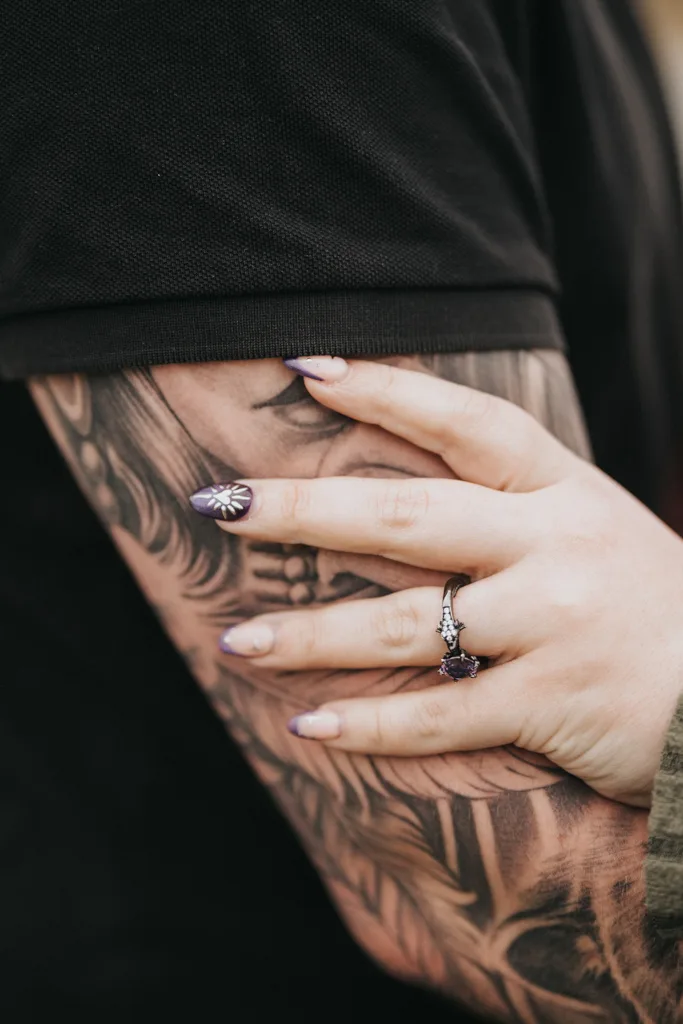 A close-up of a couples moment in Cleethorpes: one person’s tattooed arm in a black shirt, while another’s hand—with light purple nails, a silver floral ring, and an engagement ring—gently rests on it, highlighting the rings and detailed tattoo. © Aimee Lince Photography - Wedding photographer in Lincolnshire, Yorkshire & Nottinghamshire