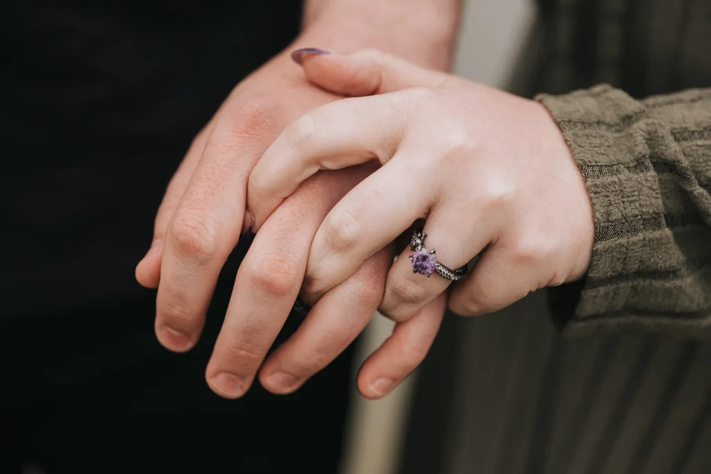 Close-up photography of two hands gently touching—one with a purple gemstone engagement ring and matching polish, the other with a darker skin tone. Both couples are dressed in long sleeves, capturing an intimate moment. © Aimee Lince Photography - Wedding photographer in Lincolnshire, Yorkshire & Nottinghamshire