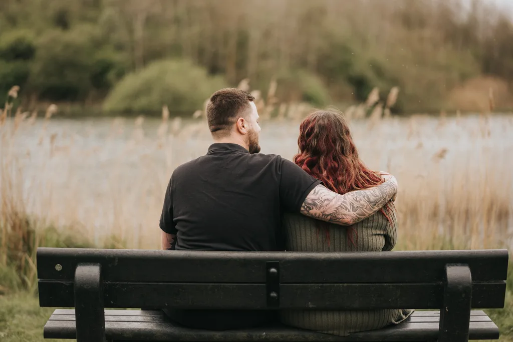 A couple sits close together on a black bench by a lake in Cleethorpes, perfect for engagement photography. The man with tattoos and a beard wears a dark shirt, while the woman in a green sweater has long, wavy, reddish hair. Tall grass and trees frame them. © Aimee Lince Photography - Wedding photographer in Lincolnshire, Yorkshire & Nottinghamshire