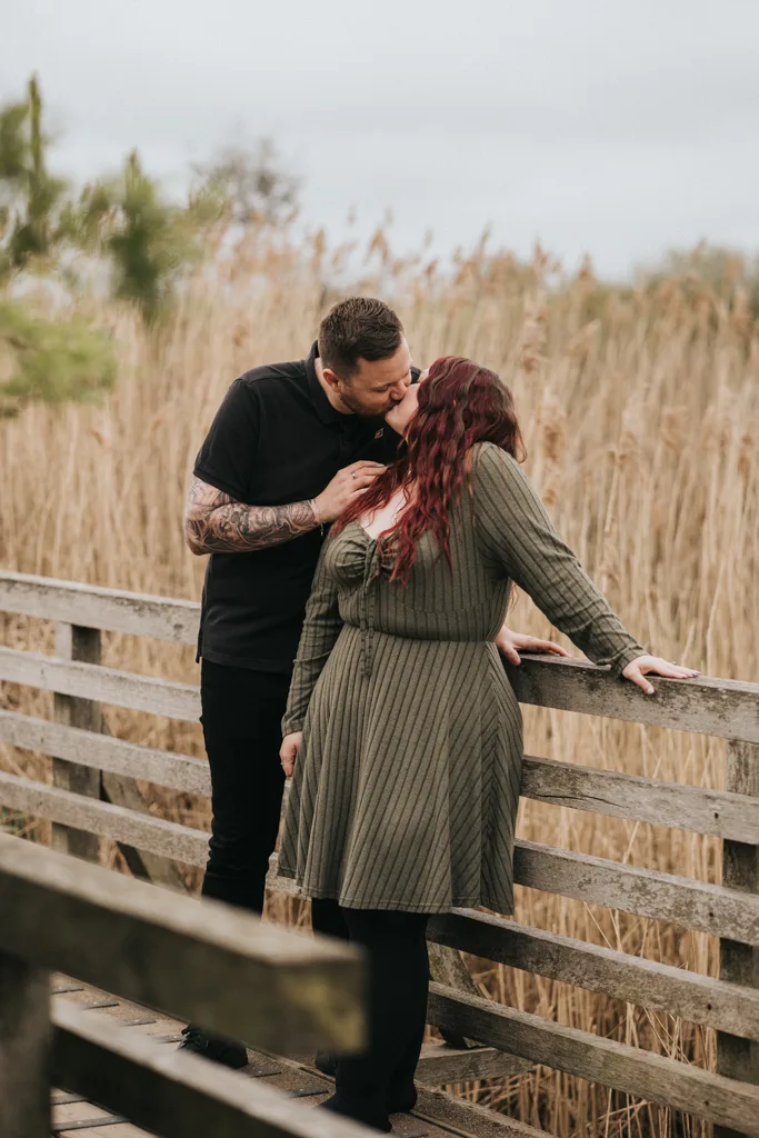 A tattooed man kisses his partner's forehead as she leans against a wooden bridge railing. This couples photography captures their tender moment outdoors, framed by tall dry grasses and a cloudy sky, both dressed in dark, casual clothes. © Aimee Lince Photography - Wedding photographer in Lincolnshire, Yorkshire & Nottinghamshire