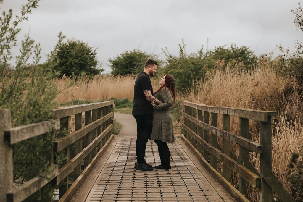 Emily & Billy Engagement Couples Phtooshoot in Cleethorpes Country Park 30 of 45 © Aimee Lince Photography - 23.04.2025 A couple stands close together on a wooden footbridge in Cleethorpes, gazing into each other’s eyes and smiling. Tall grass and trees surround them under a cloudy sky, creating a serene, intimate atmosphere perfect for an engagement moment. © Aimee Lince Photography - Wedding photographer in Lincolnshire, Yorkshire & Nottinghamshire