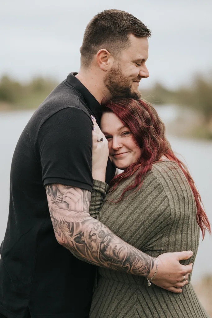 A smiling man with a beard embraces a woman with long red hair during their engagement photography in Cleethorpes. He has tattoos on his arm and wears a black shirt, while she, in a green sweater, smiles softly near water with greenery blurred behind them. © Aimee Lince Photography - Wedding photographer in Lincolnshire, Yorkshire & Nottinghamshire