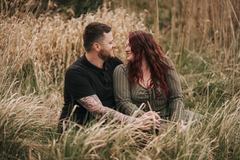 A happy couple sits closely together in tall grass, smiling at each other. With visible tattoos and a green dress, they share an affectionate moment—perfect for engagement photography in a beautiful outdoor setting. © Aimee Lince Photography - Wedding photographer in Lincolnshire, Yorkshire & Nottinghamshire