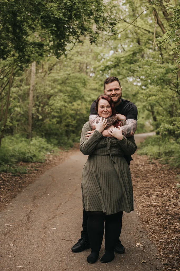 A man stands on a wooded path, smiling and hugging his partner from behind. This sweet moment of couples engagement photography is set among lush green trees on a quiet trail, with leaves scattered along the ground. © Aimee Lince Photography - Wedding photographer in Lincolnshire, Yorkshire & Nottinghamshire